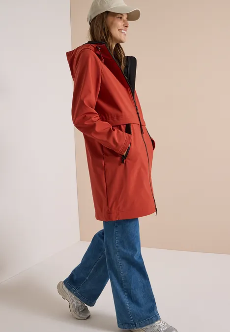 Frau trägt roten Parka mit Kapuze, Jeans, weiße Sneaker und Baseballcap, seitlich lächelnd.