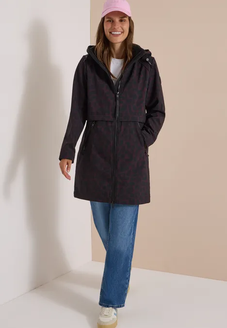 Femme souriante en imperméable à motifs avec capuche, jean, baskets rouges et casquette rose.
