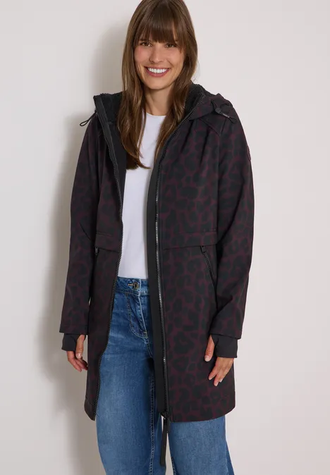 Femme souriante en manteau long rouge foncé à motifs avec capuche, t-shirt blanc et jean.