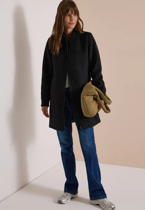 Frau in schwarzem Mantel, blauen Jeans und silbernen Schuhen hält eine khakigrüne Tasche vor hellem Hintergrund.