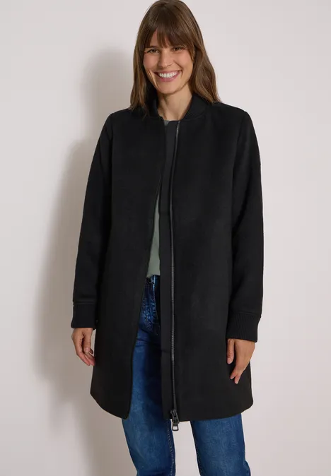 Frau in langer dunkelgrauer Jacke, Jeans und grünem Oberteil steht lächelnd vor heller Wand.