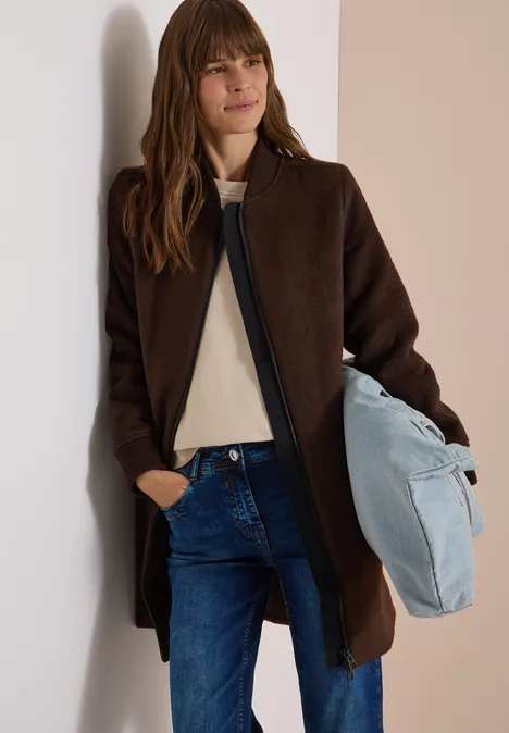 Eine Frau in braunem Mantel und Jeans lehnt an einer Wand, hält eine hellblaue Tasche.
