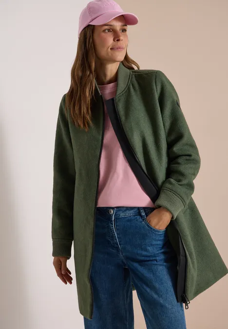 Eine Frau trägt rosa T-Shirt, grüne Jacke, Jeans und rosa Cap, steht entspannt.