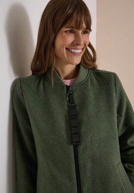Frau lächelt in grüner Jacke mit Reißverschluss und rosa Top, leicht an eine Wand gelehnt.