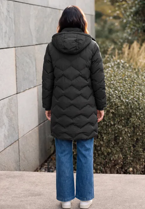 Langer Mantel mit Steppmuster Black Rückansicht einer Person in langer schwarzer Steppjacke mit Kapuze und weiter blauer Jeans vor Mauer und Sträuchern.
