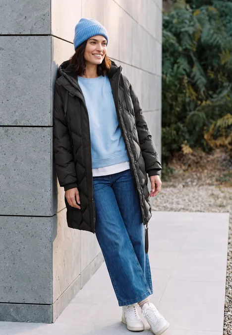 Langer Mantel mit Steppmuster Black Eine lächelnde Frau in schwarzer Winterjacke, blauer Mütze und Jeans lehnt entspannt an einer Hauswand.