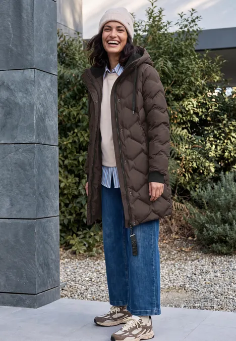 Langer Mantel mit Steppmuster chocolate sauce brown Frau in langem, gesteppter Wintermantel, Jeans und Mütze lächelt im Herbstgarten.