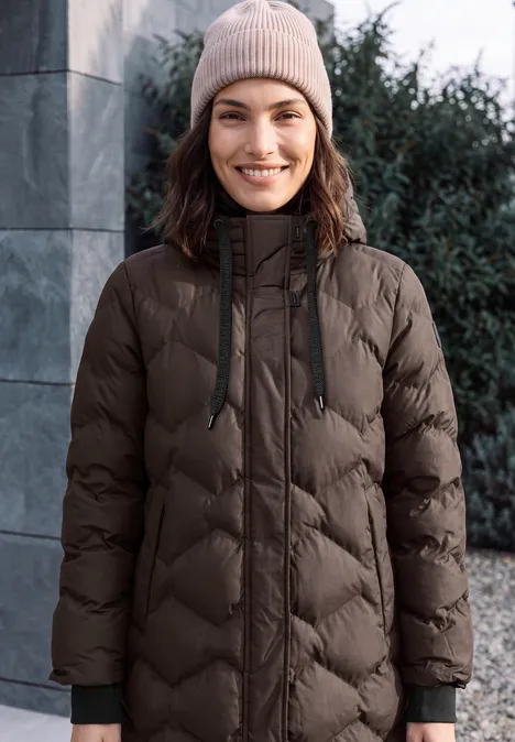 Langer Mantel mit Steppmuster chocolate sauce brown Eine junge Frau in warmer Winterjacke und Mütze lächelt im Freien in winterlicher Atmosphäre.