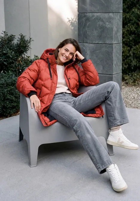 Langer Mantel mit Steppmuster light russet red Frau in roter Winterjacke und grauer Jeans sitzt entspannt auf Stuhl und lächelt.