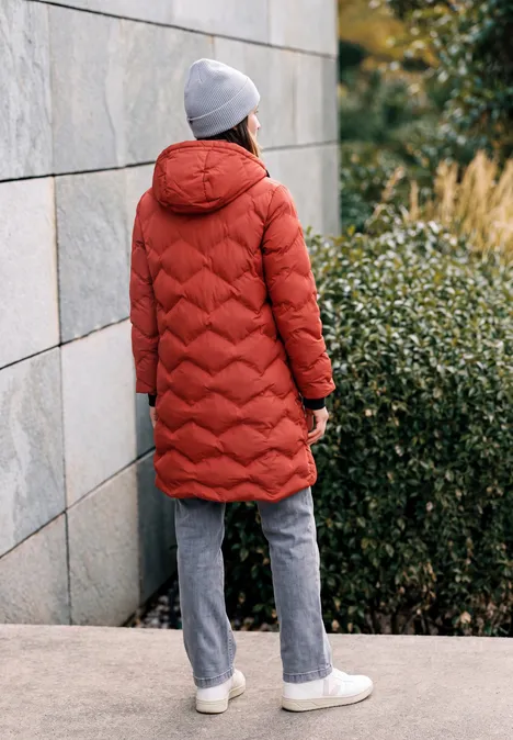 Langer Mantel mit Steppmuster light russet red Person in rotem Steppmantel, grauer Mütze, grauer Hose und weißen Sneakern steht vor einer Mauer und Büschen.