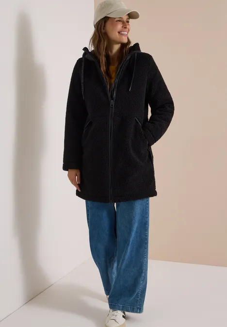 Strukturmix Teddy-Mantel mit Kapuze Black Frau in schwarzer Teddyfell-Jacke, blauer weiter Jeans und Basecap lehnt an weißer Wand.