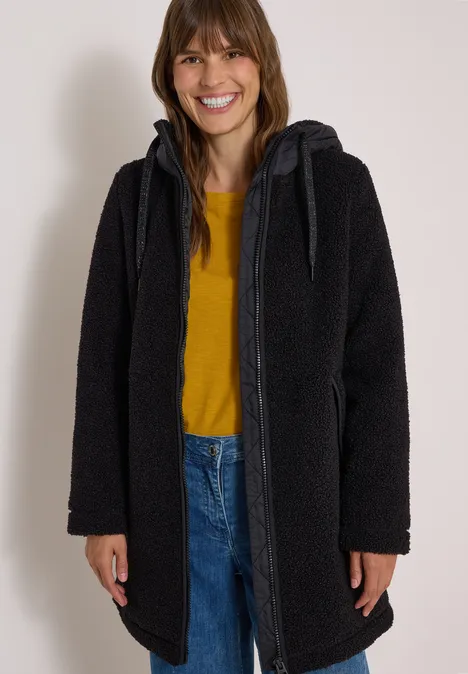 Strukturmix Teddy-Mantel mit Kapuze Black Frau in einem schwarzen Kapuzenmantel, gelbem Shirt und Jeans, die lächelt.
