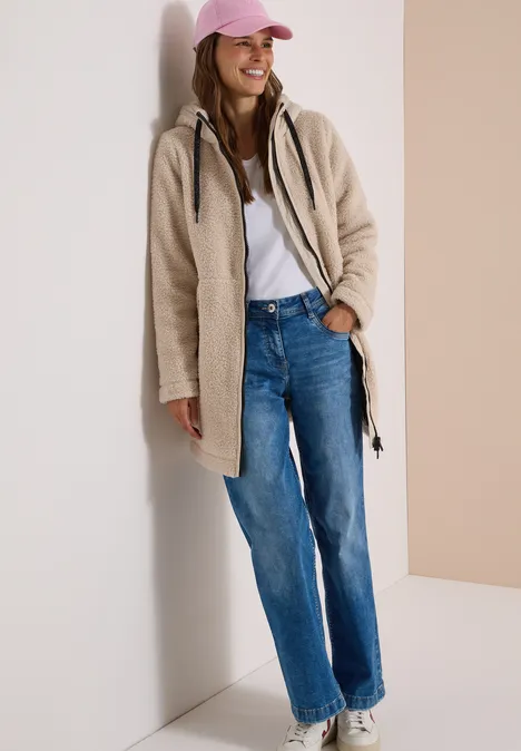 Strukturmix Teddy-Mantel mit Kapuze teddy vanilla white Eine lächelnde Frau lehnt in Mantel, Jeans und rosa Cap an einer Wand.