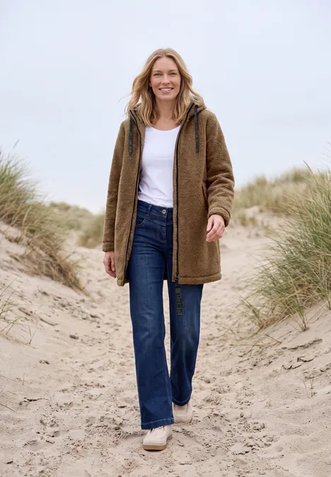 Frau in brauner Jacke, Jeans und Sneakern spaziert lächelnd über Dünensand mit Strandgras.