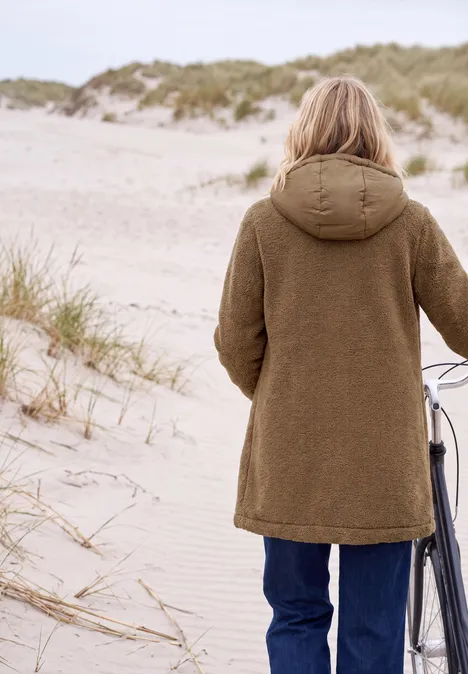 Person in braunem Mantel schiebt Fahrrad über sandige Dünen am Strand.