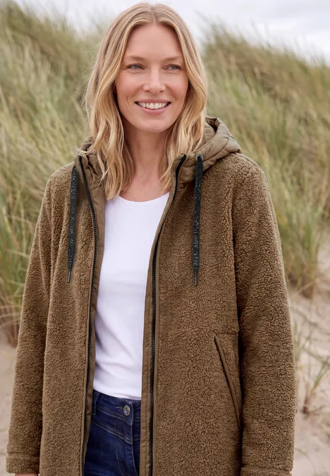 Frau mit brauner Kuscheljacke und Jeans steht vor Dünen und lächelt entspannt.