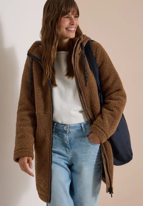 Strukturmix Teddy-Mantel mit Kapuze dark toffee brown Eine lächelnde Frau trägt eine braune Teddyjacke, weißen Pullover, blaue Jeans und eine dunkelblaue Tasche.
