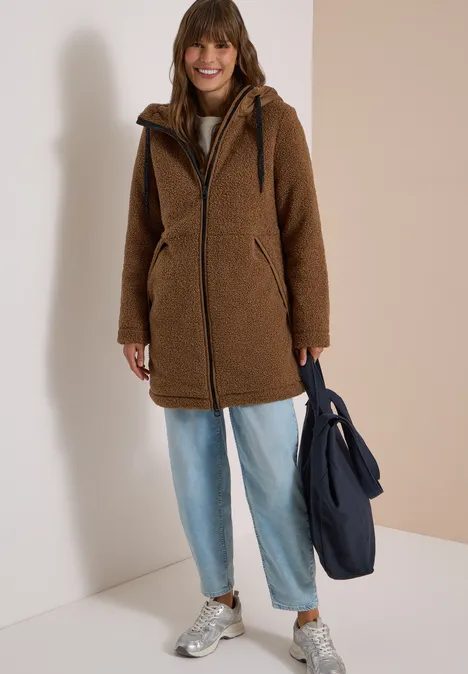 Strukturmix Teddy-Mantel mit Kapuze dark toffee brown Frau in brauner Teddyjacke, Jeans und Sneakern mit dunkelblauer Tasche vor hellem Hintergrund.