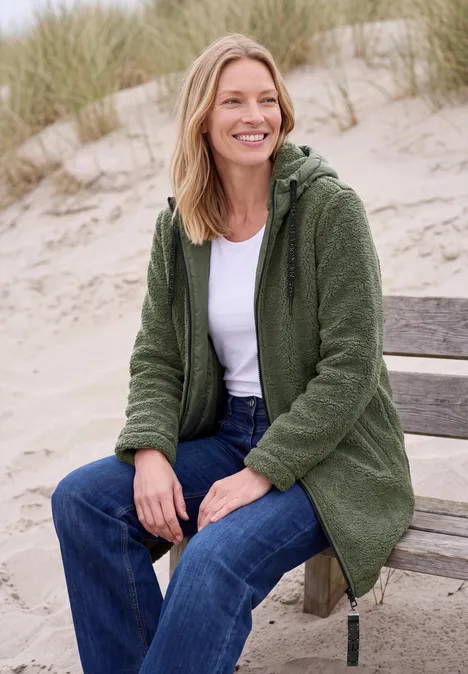 Eine lächelnde Frau in grünem Fleece-Mantel sitzt auf einer Bank am Strand vor Dünengras.