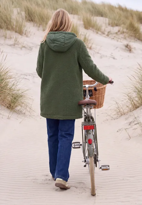 Frau in grünem Mantel schiebt ihr Fahrrad mit Weidenkorb über einen sandigen Dünenpfad.