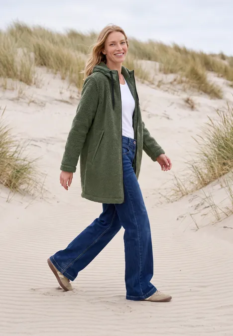 Eine Frau in grünem Mantel und Jeans spaziert lächelnd über sandige Dünen am Meer.