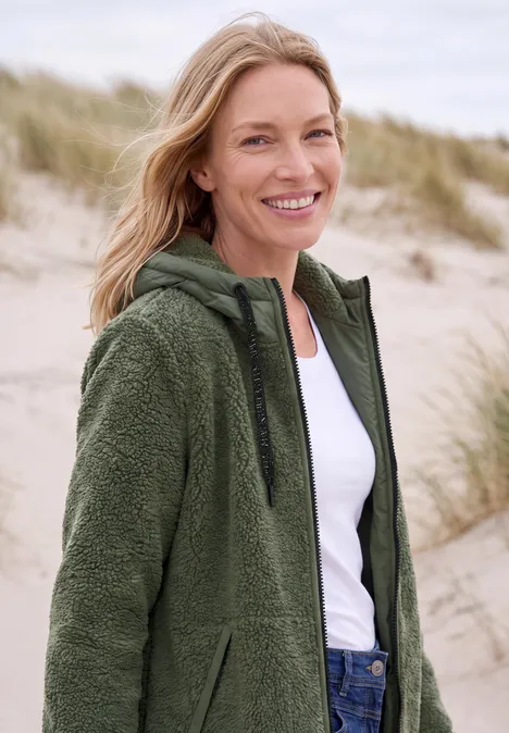 Lächelnde Frau in grüner Fleecejacke steht am Strand vor Sanddünen.
