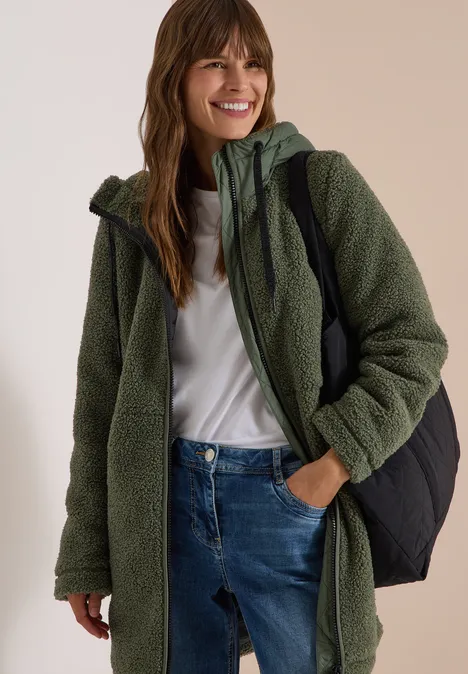 Strukturmix Teddy-Mantel mit Kapuze shadow khaki Junge Frau lächelt, trägt grüne Fleecejacke mit Kapuze, weißes Shirt, blaue Jeans und schwarzen Rucksack.