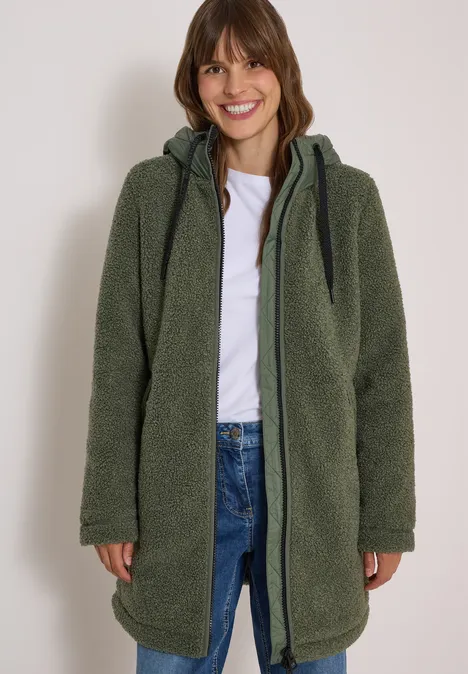 Strukturmix Teddy-Mantel mit Kapuze shadow khaki Frau mit grünem Teddy-Fleece-Mantel, Kapuze, weißem Shirt und Jeans, lächelnd.