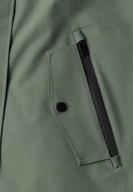 Close-up einer grünen Jackentasche mit schwarzem Reißverschluss und Druckknopf.