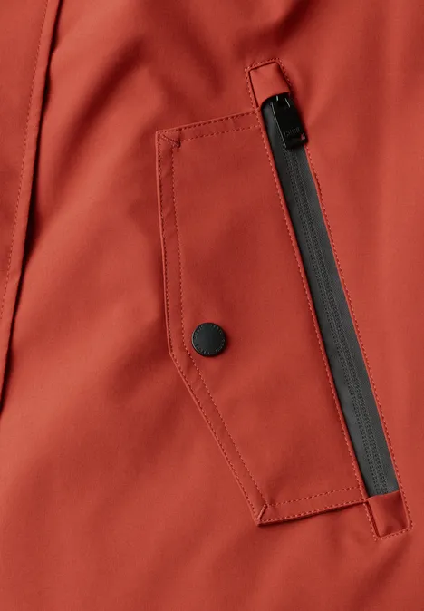 Nahaufnahme einer rostroten Jacke mit schrägem schwarzen Reißverschluss und Druckknopf an der Tasche.