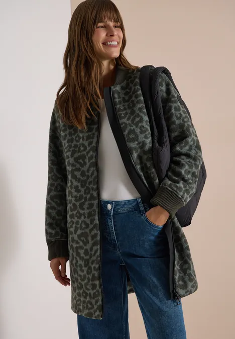 Langer Blouson mit Wollanteil olive night Frau in langer, grün-grauer Leo-Muster-Jacke mit Kapuze, weißem Oberteil, blauen Jeans und schwarzer Tasche.