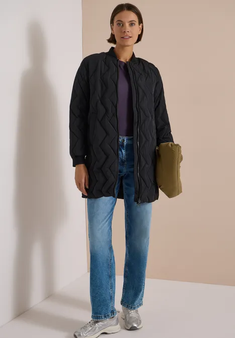 Frau in langer, gesteppter Jacke, lila Shirt, blauen Jeans und Sneakern, hält eine grüne Tasche.