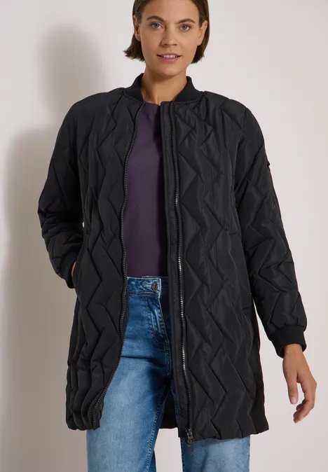 Frau in langer, gesteppter schwarzer Jacke über lila Shirt und blauen Jeans, steht mit den Händen in den Taschen.