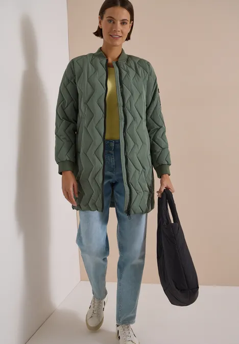 Eine Frau trägt einen gestepptern, knielangen grünen Mantel, Jeans, weiße Sneaker und hält eine schwarze Tasche.