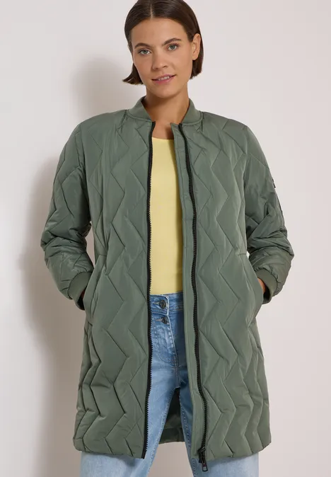 Eine Frau in einem langen, olivgrünen, gesteppten Mantel mit Zickzack-Design, gelbem Top und Jeans.