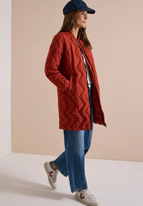 Frau mit rotem Steppmantel, blauer Jeans, weißen Sneakern und Cap vor neutralem Hintergrund.