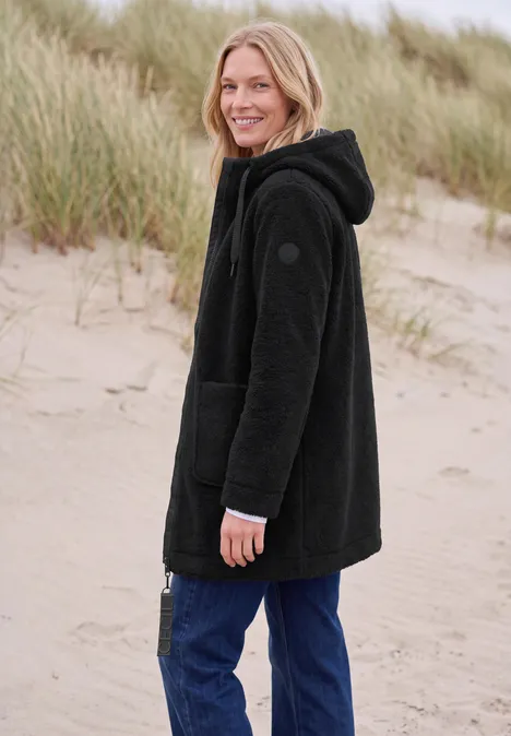 Mantel in angerauter Wolloptik Black Frau in schwarzer Fleecejacke und Jeans lächelnd am Strand vor Dünen.