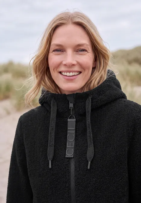 Mantel in angerauter Wolloptik Black Eine Frau lächelt in einem schwarzen Kapuzenpullover am Strand.