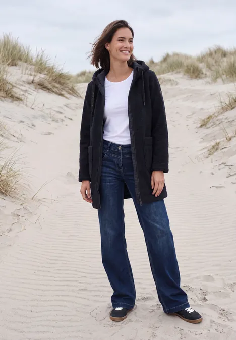 Mantel in angerauter Wolloptik dark navy blue Frau in Jeans, weißem T-Shirt und schwarzer Jacke lächelt, steht auf sandigen Dünen mit Strandhafer.