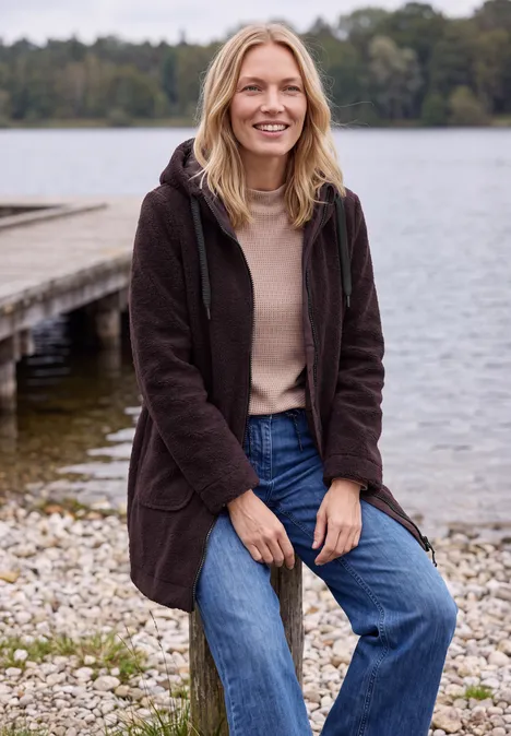 Jas met een ruwe wollook berry red Een vrouw in een bruine jas zit ontspannen op stenen aan de oever van een meer, met een steiger op de achtergrond.