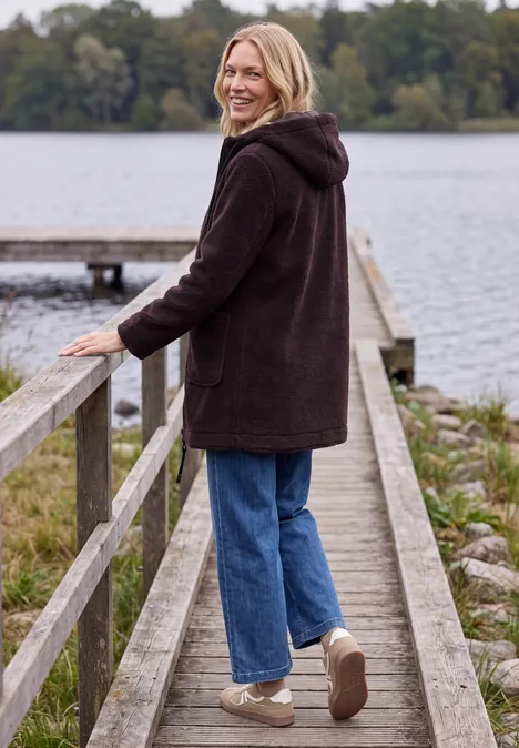 Jas met een ruwe wollook berry red Vrouw in donker mantel op steiger bij meer, kijkt lachend om, met bomen op de achtergrond.