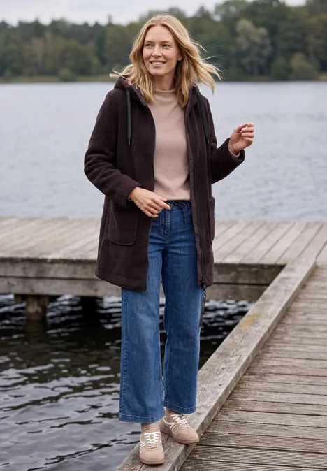 Jas met een ruwe wollook berry red Vrouw in jas, jeans en sneakers wandelt glimlachend over een steiger bij een meer.