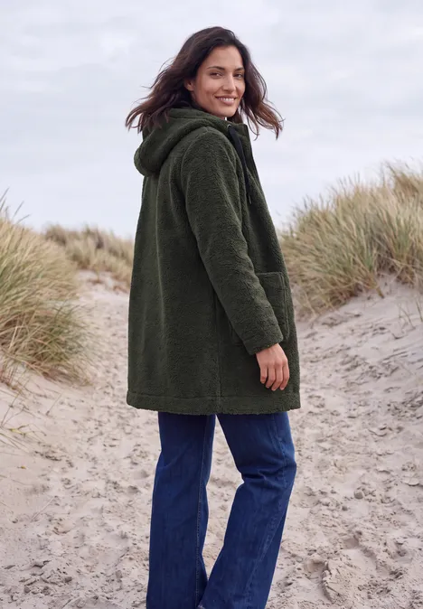 Mantel in angerauter Wolloptik olive night Eine Frau steht auf einem Dünenpfad am Strand, trägt eine grüne Jacke und schaut über die Schulter.