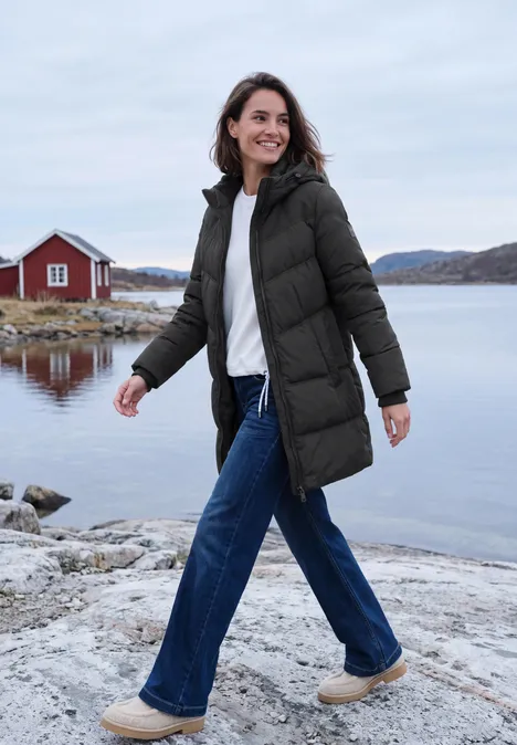 Eine lächelnde Frau in grüner Winterjacke spaziert an einem felsigen Ufer mit Wasser.