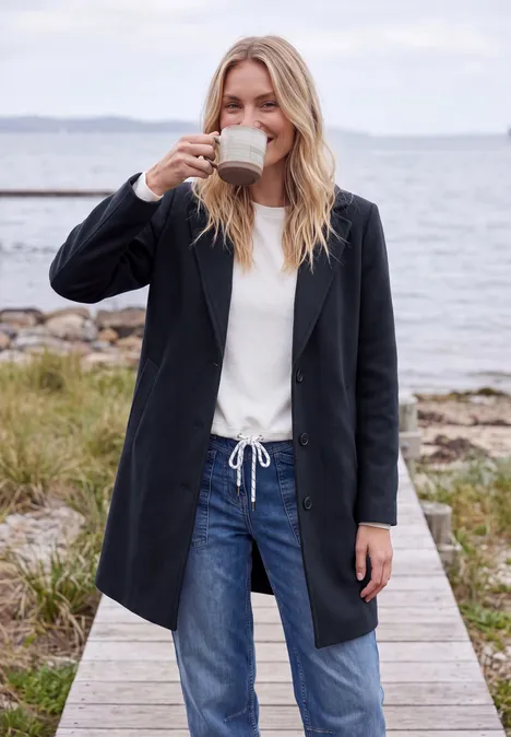 Manteau à revers aspect laine dark navy blue Frau im schwarzen Mantel trinkt Kaffee auf einem Steg, Jeans und weißes Shirt, Meer im Hintergrund.