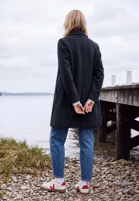 Manteau à revers aspect laine dark navy blue Eine Person steht an einem steinigen Ufer und blickt auf das Wasser, trägt Mantel und Jeans.