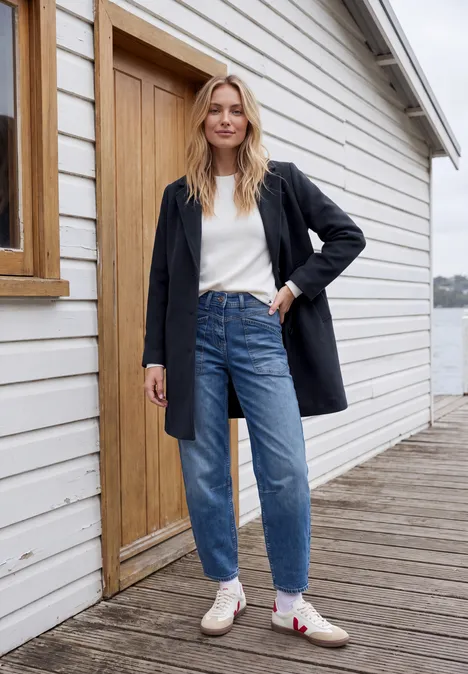 Manteau à revers aspect laine dark navy blue Eine Frau in Jeans, weißem Pullover, schwarzem Mantel und weißen Stiefeln steht auf einem Holzsteg vor einer weißen Hütte.