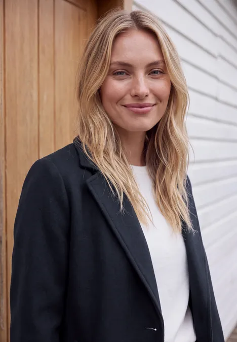Manteau à revers aspect laine dark navy blue Lächelnde Frau mit blondem Haar, schwarzem Blazer und weißem Shirt lehnt vor Holz- und Wandverkleidung.