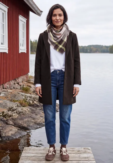Jas met revers in wollook chocolate sauce brown Eine Frau im Mantel, Jeans und Schal steht auf einem Steg vor einem roten Bootshaus am See.
