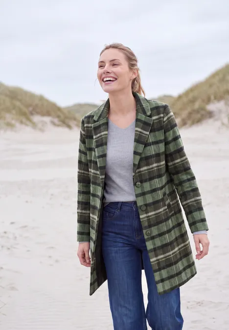 Eine lächelnde Frau in grün kariertem Mantel, grauem Shirt und Jeans am Strand vor Dünen.